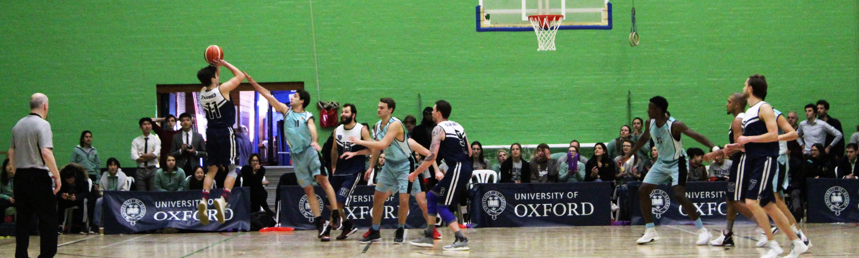 Men playing basketball in the Oxford vs Cambridge Varsity match, with a crowd watching from behind the basket